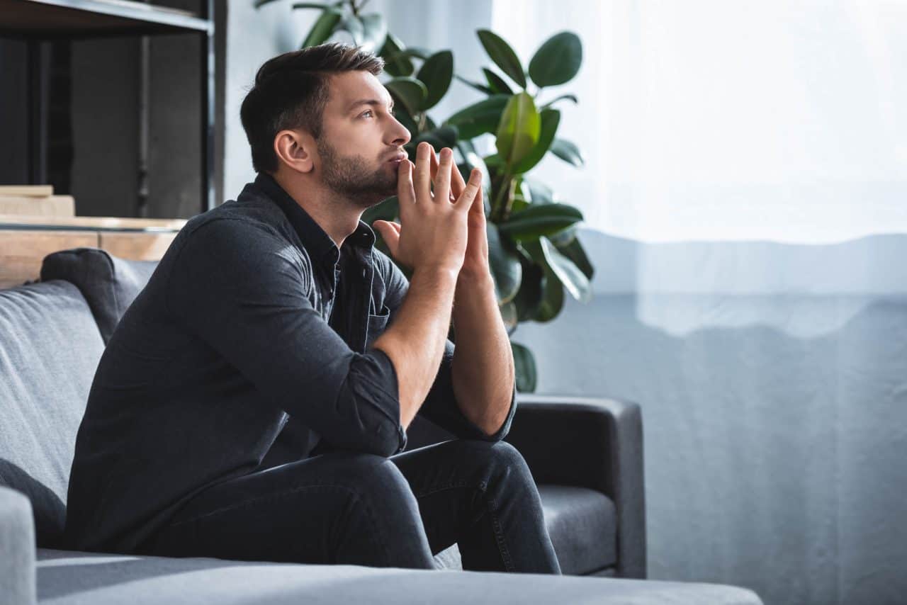 Man sitting on sofa deep in thought with hands clasped near face.