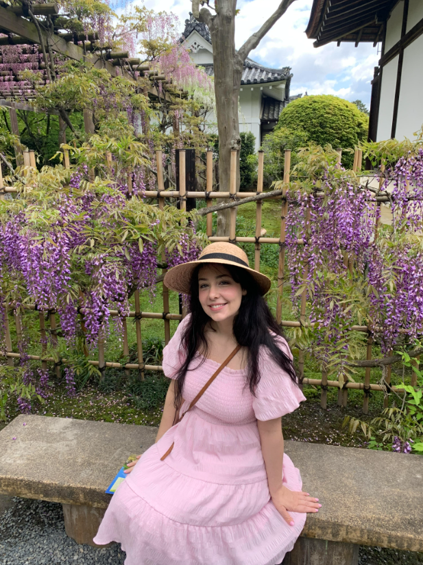 Woman in pink dress and straw hat sitting on stone bench in garden with purple flowers.