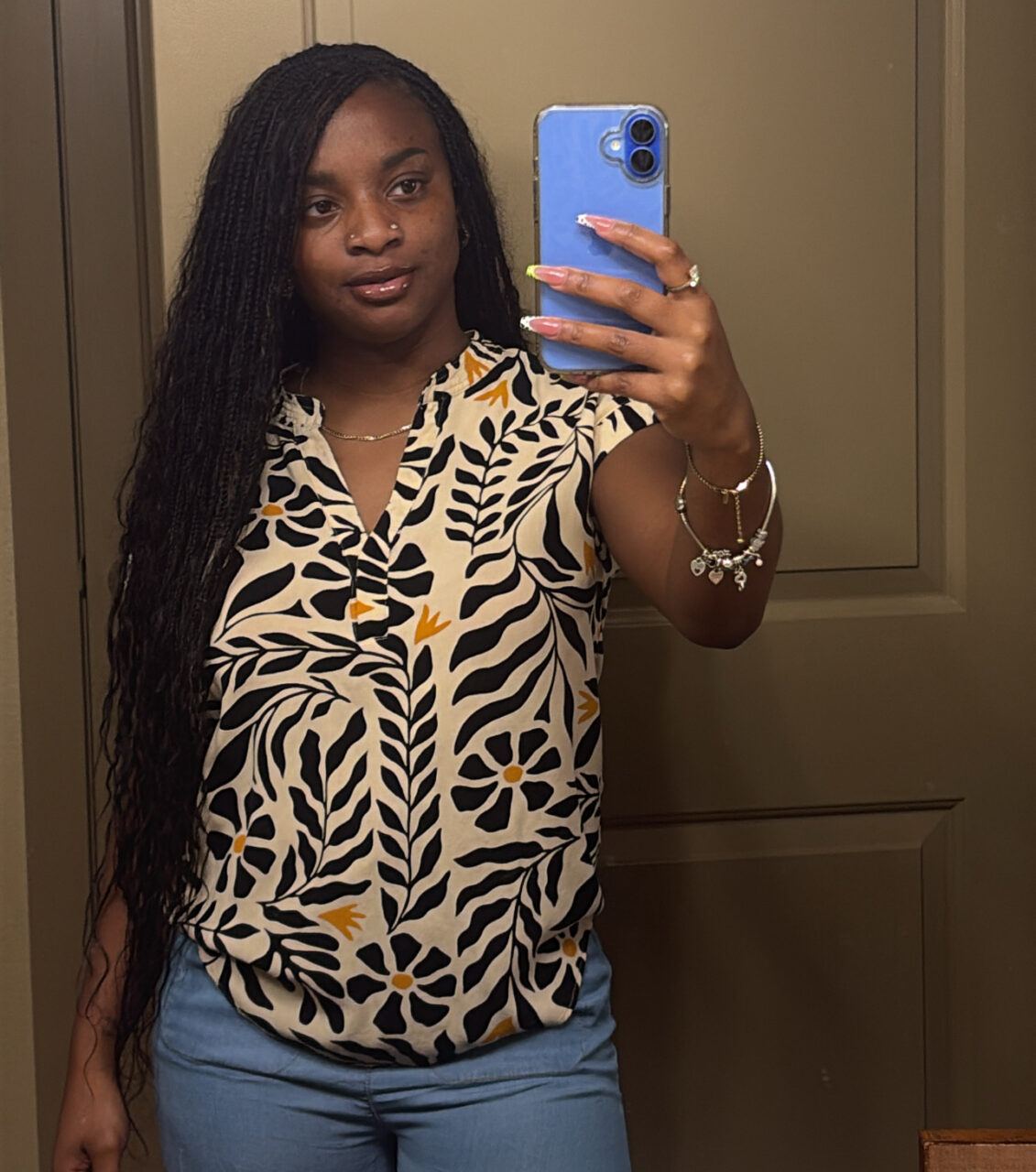 Woman taking a mirror selfie with a blue phone, wearing a patterned blouse and jeans.