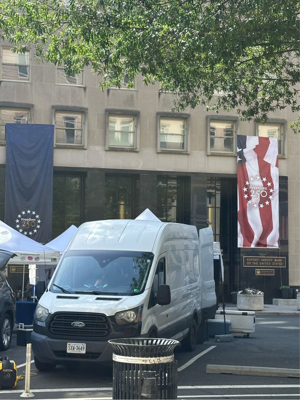 A white delivery van parked in front of a building with patriotic 250th anniversary banners.