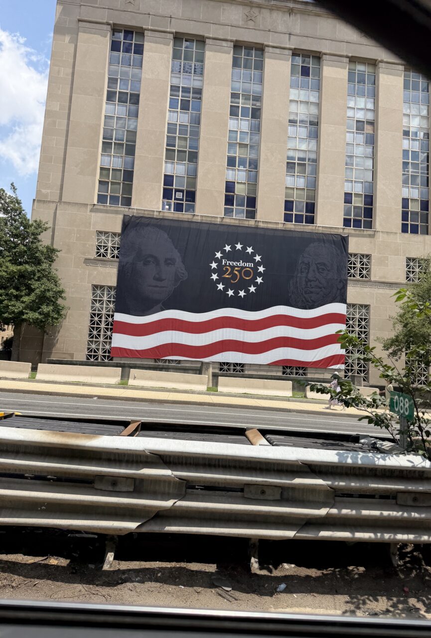 Billboard celebrating 250th Independence Day with American flag design and portraits.