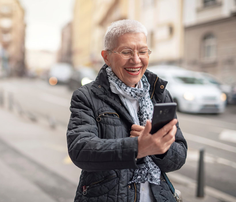 Elderly woman smiling while using her smartphone outdoors in an urban setting.